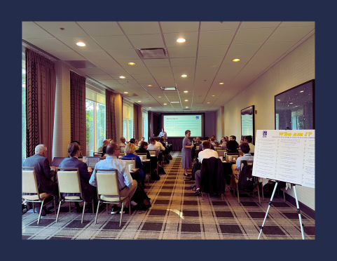 Presenter speaks to attendees in a conference room with a slide on screen and a 'Who am I?' poster in view